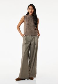 Brauner gestrickter ärmelloser Pullover mit strukturiertem Muster, kombiniert mit hoch taillierten, weit geschnittenen grauen Hosen und braunen Wildlederschuhen.