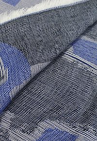 Tessuto intrecciato con un motivo geometrico in tonalità di blu e grigio, con una consistenza morbida e bordi frangiati, che mostra dettagli intricati.