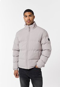 Jachetă pufoasă de culoare gri deschis cu guler înalt, fermoar frontal, manșete elastice și buzunare laterale. Are un patch cu logo negru pe mânecă.
