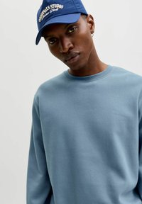 Sudadera azul claro con cuello redondo y mangas largas, hecha de un tejido suave. Se lleva con una gorra azul que presenta texto bordado.