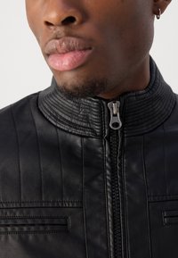 Jachetă din piele neagră cu detalii cusute pe guler și umeri, având un închidere cu fermoar frontal și accente de cusături orizontale.