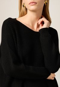 Czarny sweter o fakturowanej strukturze z szerokim dekoltem i długimi rękawami. Modelka nosi efektowne złote i czarne kolczyki, prezentujące elegancki wzór.