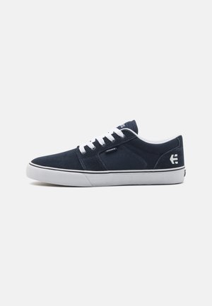 Navy blue suede sneakers with white laces, a white rubber sole, and subtle branding on the side and tongue. Smooth texture and low-top design.