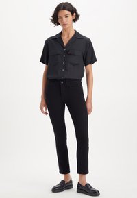 Zwarte kortarmshirt met knoopsluiting aan de voorkant en twee borstzakken, gecombineerd met zwarte aansluitende jeans en zwarte instapschoenen. Gladde stoftextuur.