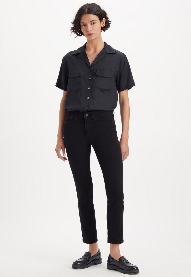 Chemise noire à manches courtes avec boutons sur le devant et deux poches à la poitrine, associée à un jean noir ajusté et des mocassins noirs. Texture de tissu lisse.