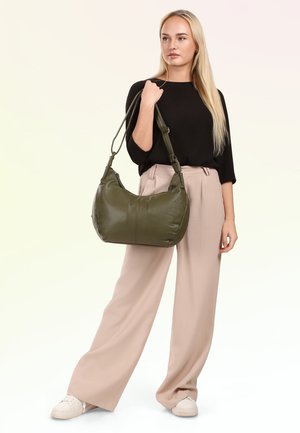 Frau trägt schwarzen Top und beigefarbene Hosen mit weitem Bein, hält große olivgrüne Umhängetasche mit Träger, steht vor weißem Hintergrund.