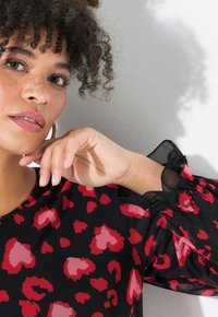 Blusa negra con estampado de leopardo rojo y rosa, con mangas abullonadas transparentes y un escote redondeado. La modelo muestra la mano colocada bajo la barbilla.