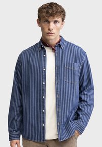 Striped denim shirt in blue and white, featuring a button-down collar, chest pocket, and relaxed fit, layered over a white crew-neck top.