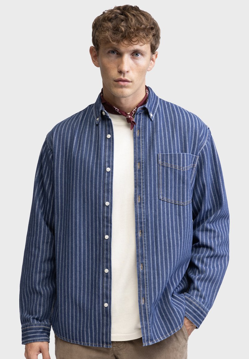 Striped denim shirt in blue and white, featuring a button-down collar, chest pocket, and relaxed fit, layered over a white crew-neck top.