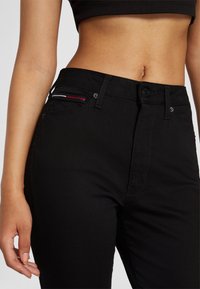 Cintura de mujer vestida con jeans de tiro alto negros que tienen un pequeño detalle de franjas rojas, blancas y azules cerca del bolsillo delantero y una camiseta recortada negra.