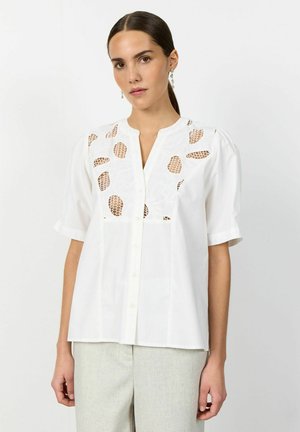 Fehér, rövid ujjú blúzt viselő nő, vállán és mellkasán kivágott csipkedíszítéssel, világosszürke nadrággal, fehér háttér előtt áll.