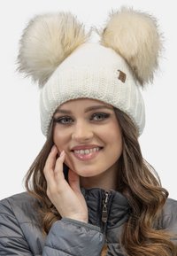 Bonnet en tricot blanc avec deux grands pompons en fausse fourrure sur le dessus. Il présente une petite étiquette de marque à l'avant et une texture côtelée.