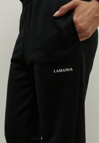 Čierne teplákové nohavice s hladkou textúrou, vybavené elastickým pásom a bočnými vreckami. Na ľavom stehne je biele logo "LAMANIA".