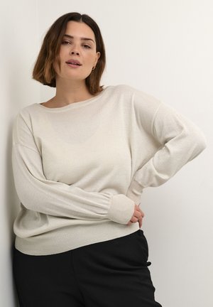 Light, metallic-knit sweater in cream, featuring a boat neckline, dropped shoulders, and snug ribbed cuffs; paired with black trousers.