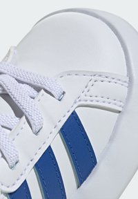 Scarpa sportiva bianca realizzata in pelle sintetica, con strisce blu, lacci bianchi e dettagli cuciti. Texture liscia e punta arrotondata.