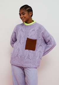 Persona sorridente con capelli intrecciati che indossa un maglione lavorato a maglia grosso color lavanda con tasca marrone, scollo giallo neon e pantaloni coordinati.