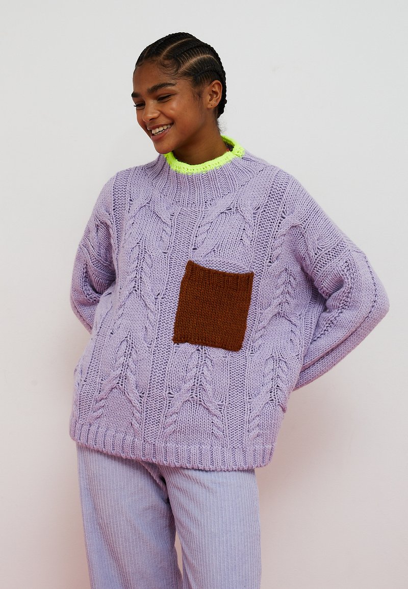 Persona sorridente con capelli intrecciati che indossa un maglione lavorato a maglia grosso color lavanda con tasca marrone, scollo giallo neon e pantaloni coordinati.