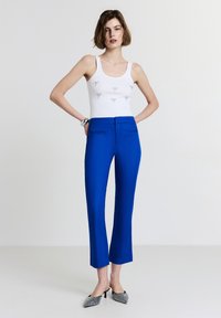 Top bianco senza maniche con ricami di fiocchi, abbinato a pantaloni su misura blu brillante. La modella indossa scarpe con tacchi a pois bianchi e neri.