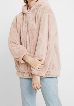 Person iført en lyserød, fluffy lynlåshættejakke med elastiske manchetter og kant, der holder den ene arm, iført lyseblå jeans.