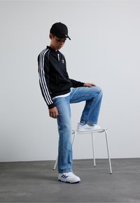 Czarna sportowa kurtka z białymi paskami, jasnoniebieskie dżinsy i białe sneakersy z niebieskimi akcentami. Model siedzi na białym krześle.