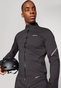 Schwarze wasserdichte Radjacke mit hohem Kragen, Frontreißverschluss und reflektierenden Akzenten. In der anderen Hand wird ein schwarzer Helm gehalten.