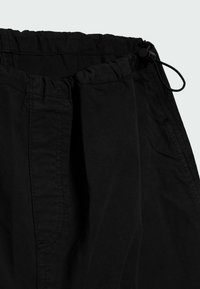 Schwarze, leichte Hose mit elastischem Bund und Kordelzug. Verfügt über subtile Textur und seitliche Taschen. Nahaufnahme des oberen Bereichs.