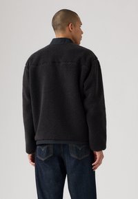 Sort fleecejakke med en blød, tekstureret overflade og en struktureret krave. Båret over mørke denim jeans, der viser et lige snit.