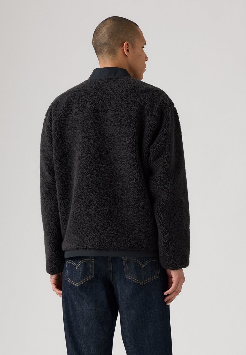 Zwarte fleece jas met een zachte, gestructureerde oppervlakte en een gestikte kraag. Draagt over een donkere spijkerbroek, die een recht gesneden ontwerp laat zien.