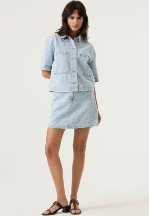 Veste en denim bleu clair à manches courtes, présentant un motif cachemire texturé, assortie à une jupe en denim assortie. Sandales à brides marron.