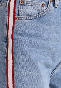 Vaqueros de mezclilla de un azul claro con una franja lateral en rojo y blanco, con remaches de cobre y detalles de costura cerca de los bolsillos.