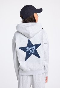 Grijze hoodie met een capuchon met trekkoord, voorzien van een grote donkerblauwe ster en de tekst "Lucky Me" op de achterkant. Zachte textuur, casual pasvorm.
