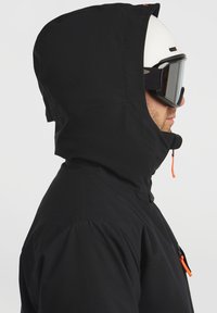 Zwarte buitenjack met hoge kraag en bevestigde capuchon. Voorzien van een oranje trekkoord en een gladde, matte textuur. Zwarte skibril wordt gedragen.