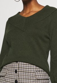 Femme portant un pull en maille vert foncé avec un large col en V à côtes, associé à une jupe taille haute en pied-de-poule, ornée de boutons et dotée de poches avant.