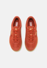 Red suede sneakers with a rounded toe, textured surface, and contrasting gum rubber sole. Features flat laces and logo accents.