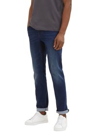 Jeans de mezclilla azul oscuro con corte recto, acentos descoloridos y puños enrollados, combinados con zapatillas blancas, usados con una camiseta gris.