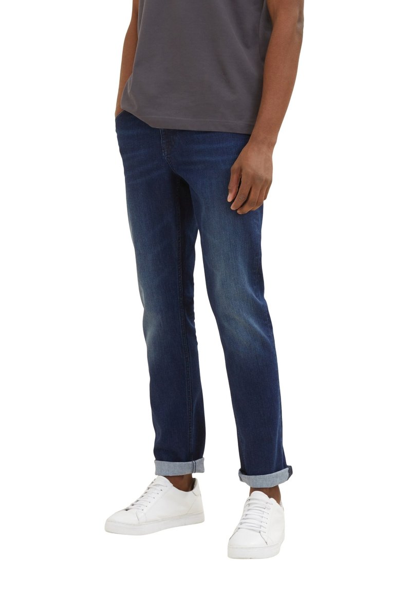 Jeans de mezclilla azul oscuro con corte recto, acentos descoloridos y puños enrollados, combinados con zapatillas blancas, usados con una camiseta gris.