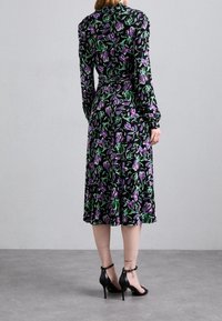 Černé šaty s dlouhým rukávem a květinovým vzorem v purpurové a zelené barvě, detail s páskem v pase a rozšířená sukně; nošené s černými podpatky.