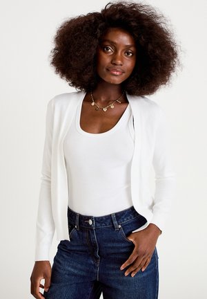 Kobieta o naturalnie kręconych włosach, ubrana w biały kardigan, biały top na ramiączkach, ciemnoniebieskie dżinsy oraz złoty naszyjnik, z dłonią w kieszeni.