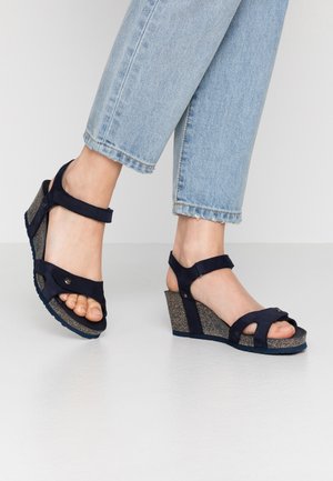 Fødder iført blå kilehælssandaler med justerbare remme, kombineret med lyseblå afkortede jeans mod en ensfarvet baggrund.