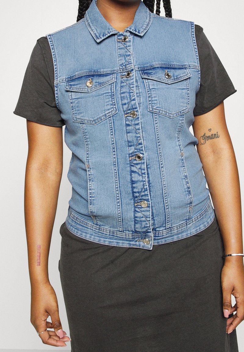 Džínová vesta v světle modré barvě, se límcem, předními kapsami a zapínáním na knoflíky. Nositá přes tmavé tričko, zvýrazňující kontrast barev.