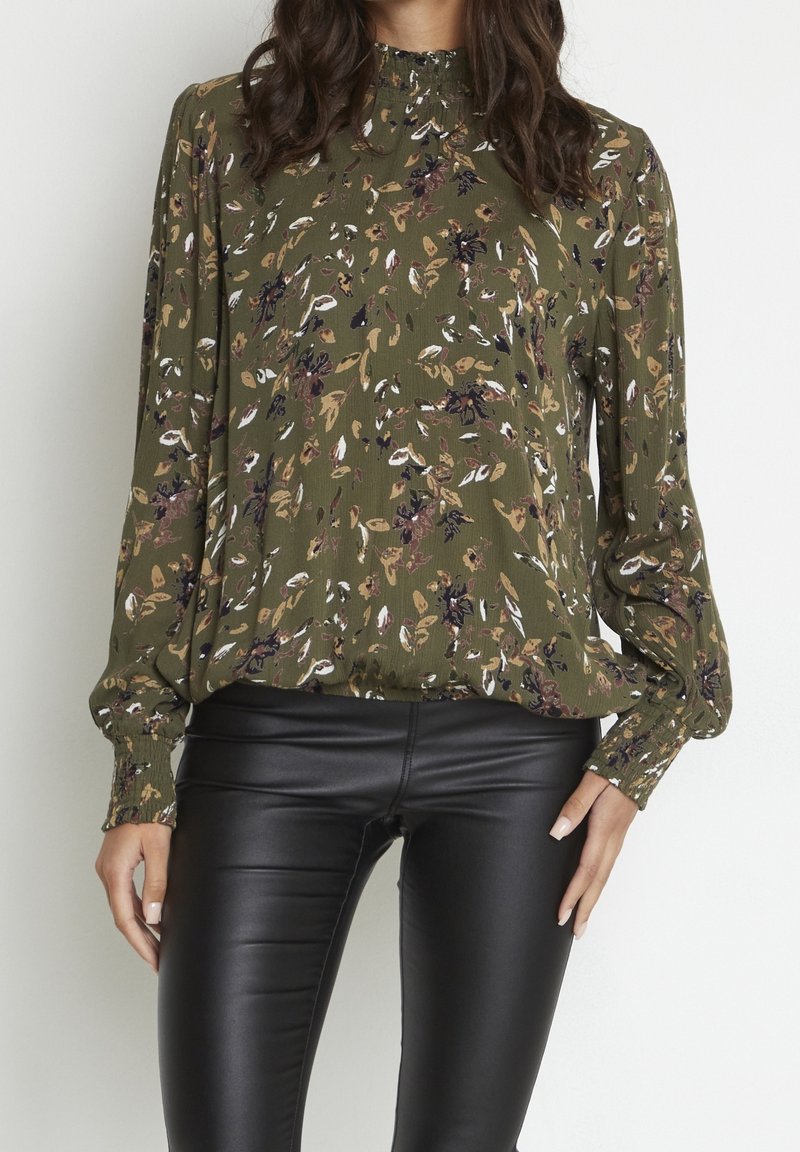 Blouse à fleurs verte en tissu léger, avec manches longues, un col plissé et des accents noirs. Associée à des leggings noirs brillants.