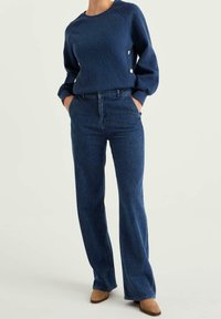 Marinblå texturerad cardigan med knappar på sidan, kombinerad med mörkblå utsvängda jeans. Båda plaggen har en avslappnad passform och enkel design.