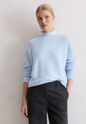 Pull en tricot bleu clair à col montant et manches longues, associé à un pantalon noir. Tissu texturé avec une coupe décontractée.