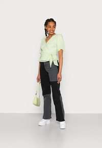 Top envolvente texturizado verde claro com mangas bufantes, emparelhado com calças em patchwork preto e cinza. Segurando uma pequena bolsa verde claro, vestindo sapatos plataforma brancos.