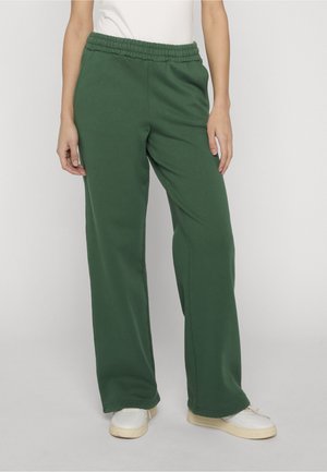 Donna che indossa pantaloni sportivi larghi verde scuro con fascia elastica in vita e scarpe da ginnastica bianche, in piedi contro uno sfondo chiaro e uniforme.