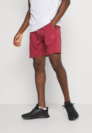 Mężczyzna ubrany w bordowe spodenki Adidas, białą koszulkę i czarne sneakersy, stoi z jedną ręką w kieszeni na białym tle.