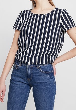 Femme portant un chemisier à manches courtes rayé verticalement bleu marine et blanc, rentré dans un jean en denim bleu taille haute.
