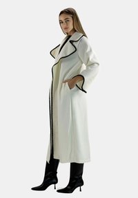 Lang hvid frakke med sort flæset kant og lommer, med brede ærmer. Kombineret med sorte knæhøje støvler. Tekstureret stof er synligt.