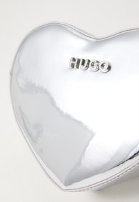 Borsa a forma di cuore argento con superficie lucida e rifiniture bianche. Presenta il logo metallico in rilievo "HUGO" sulla parte anteriore.