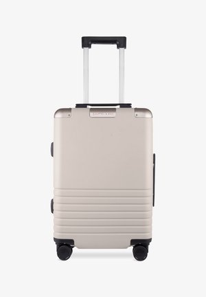 Hard-shell suitcase with a smooth beige finish, retractable black handle, and four black wheels. Notable horizontal grooves on the surface.